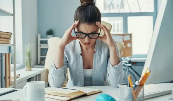 Femme stressée au bureau, mains sur les tempes devant un carnet ouvert