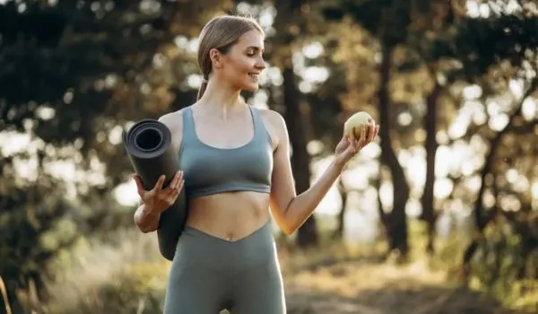 Femme en tenue de sport en plein air tenant un tapis de yoga et un fruit, symbole d’un mode de vie sain