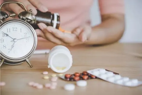 Main tenant des comprimés, avec pilulier ouvert, plaquettes de médicaments et réveil sur une table