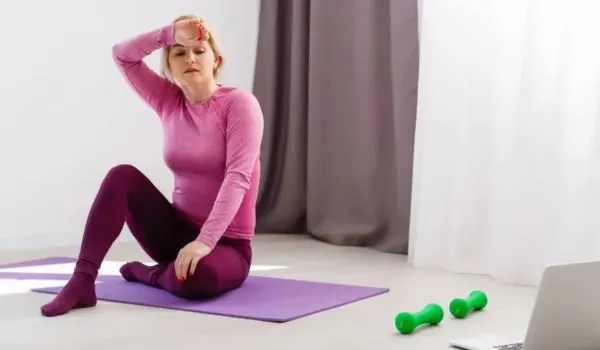 Femme assise sur un tapis de sport, l’air fatigué, avec haltères et ordinateur à proximité