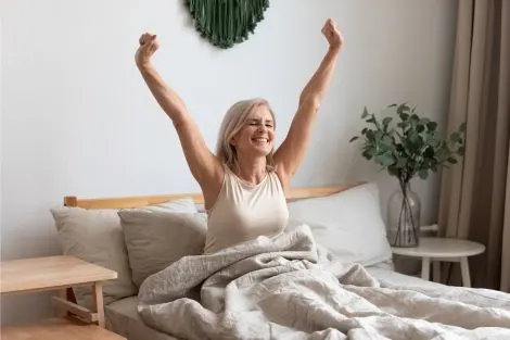 Femme senior heureuse se réveillant reposée dans un lit blanc lumineux, bras levés, après
une nuit réparatrice