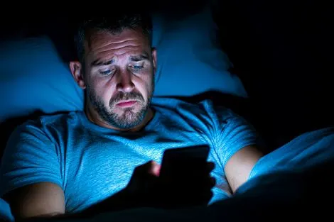 Homme allongé dans son lit regardant son téléphone dans l'obscurité, perturbant son cycle du sommeil avec la lumière bleue