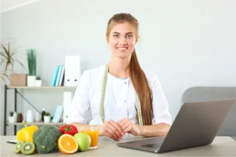 Une professionnelle de la santé souriante suit son poids avec des fruits et un ordinateur.