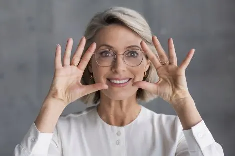 Femme d'âge mûr, cheveux blonds courts et lunettes, souriante, mains levées près du visage
