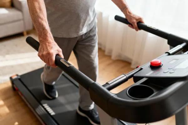 Senior marchant sur un tapis de course à domicile avec de longues barres de maintien latérales robustes. Focus sur les mains tenant la barre et la console avec un gros bouton d'arrêt d'urgence rouge visible. Posture sécurisée et confiante