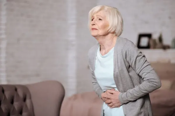 Femme senior debout, se tient le ventre avec une expression douloureuse, illustrant l'inconfort digestif et le transit intestinal ralenti fréquent avec l'âge