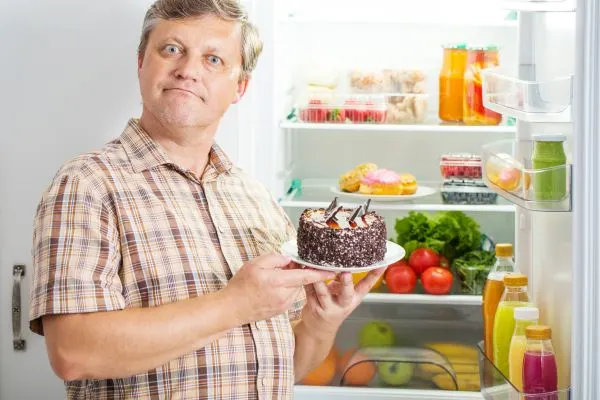 homme devant un refrigerateur ouvert dans sa main une patisserie illustrant les envies de sucre liees a la serotonine