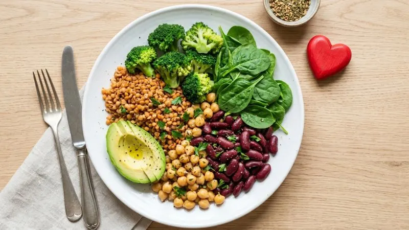 assiette vue de dessus remplie de légumineuses colorées lentilles corail pois chiches dorés haricots rouges accompagnée
de légumes verts frais brocoli épinards avocat avec un petit cœur rouge posé sur le côté symbolisant la santé cardiovasculaire.