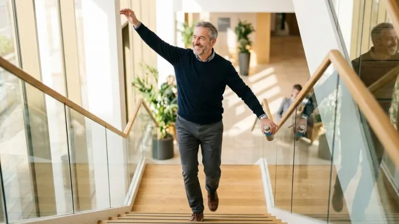 Un homme de 55 ans marchant d'un pas assuré dans un escalier et s'étirant
doucement avec un léger sourire, l'air légère et confiante.